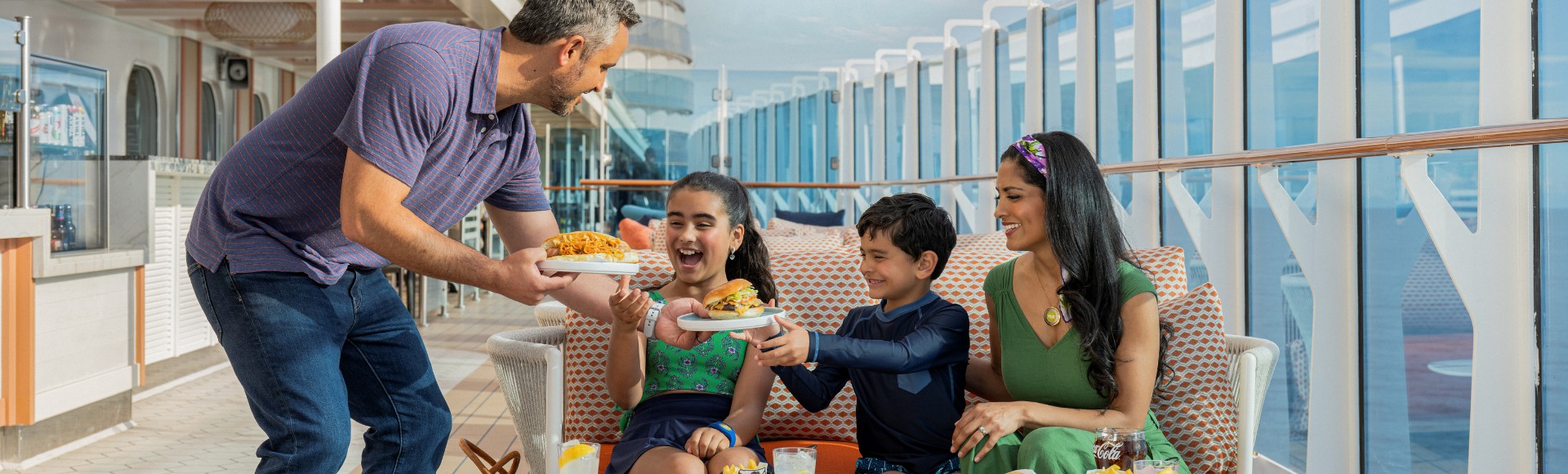 A family enjoys a meal on a cruise ship deck. A man serves fries to two smiling children seated on an orange sofa, with a woman beside them. Drinks and food are on the table. The scene is sunny and joyful.