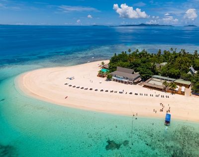 Aerial view of a tropical island with white sandy shores, lush greenery, and clear turquoise water. Beach chairs line the sand, evoking a serene vacation setting.