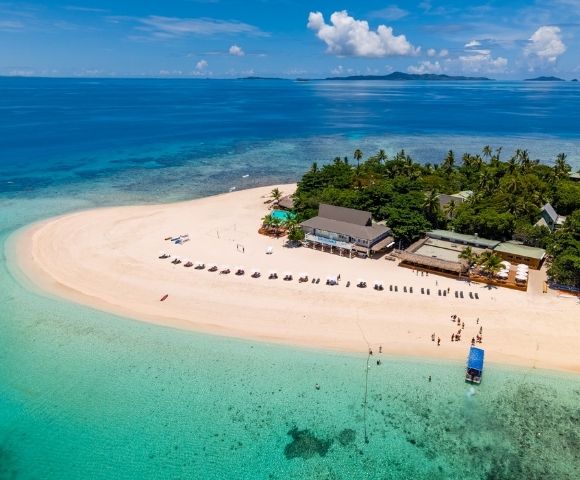Aerial view of a tropical island with white sandy shores, lush greenery, and clear turquoise water. Beach chairs line the sand, evoking a serene vacation setting.