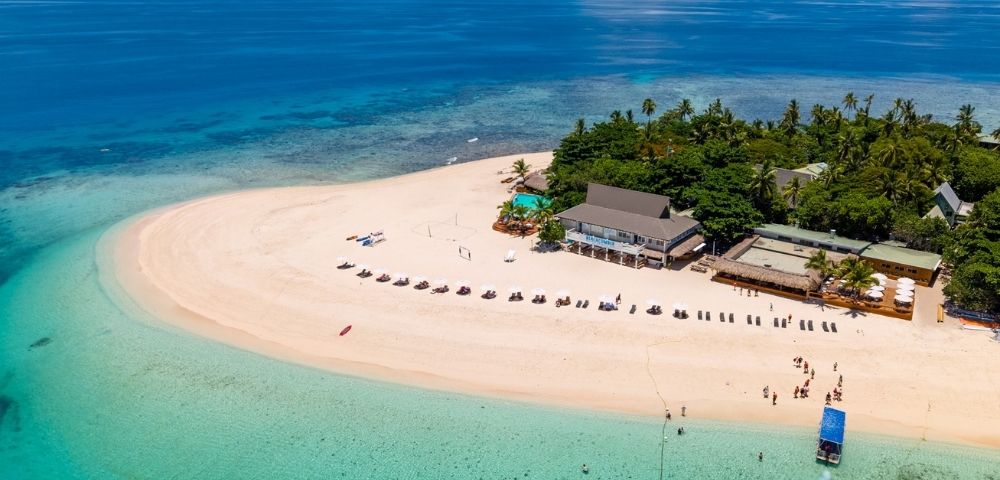 Aerial view of a tropical island with white sandy shores, lush greenery, and clear turquoise water. Beach chairs line the sand, evoking a serene vacation setting.