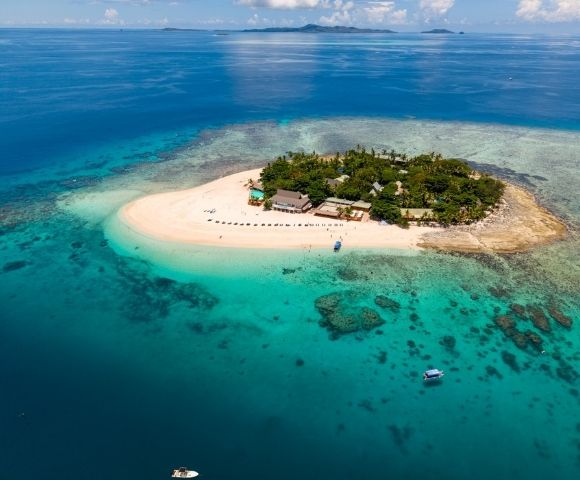 Aerial view of a small tropical island with lush greenery and sandy beaches, surrounded by turquoise water, coral reefs, and a few boats nearby.