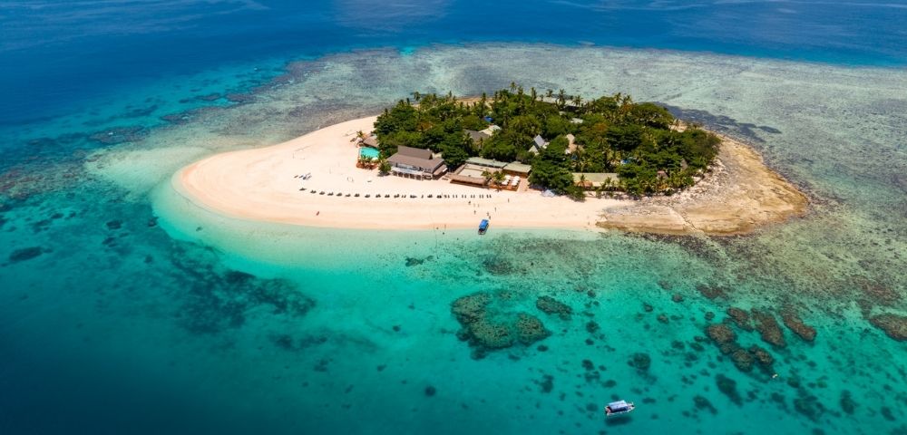 Aerial view of a small tropical island with lush greenery and sandy beaches, surrounded by turquoise water, coral reefs, and a few boats nearby.