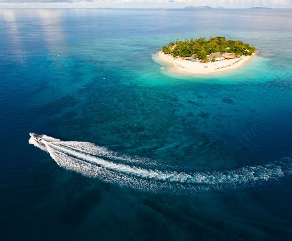 Aerial view of a speedboat leaving a white wake as it circles a small, lush tropical island surrounded by clear blue ocean under a partly cloudy sky.