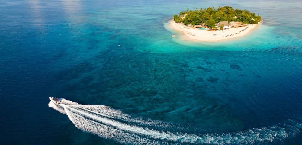 Aerial view of a speedboat leaving a white wake as it circles a small, lush tropical island surrounded by clear blue ocean under a partly cloudy sky.