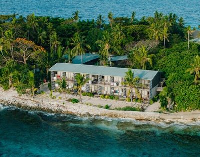 Aerial view of a coastal retreat with a long, two-story building surrounded by lush tropical trees, adjacent to a vibrant blue ocean.