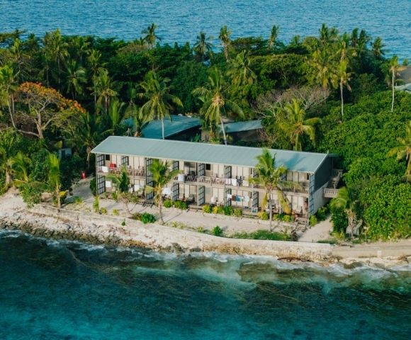 Aerial view of a coastal retreat with a long, two-story building surrounded by lush tropical trees, adjacent to a vibrant blue ocean.