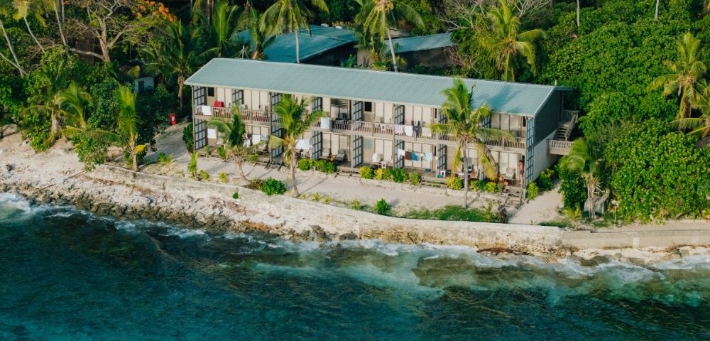 Aerial view of a coastal retreat with a long, two-story building surrounded by lush tropical trees, adjacent to a vibrant blue ocean.