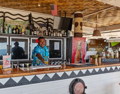 Bartender in blue floral shirt and red flower serves a tropical drink at a festive beach bar. Bottles line the counter; an American flag is visible above.