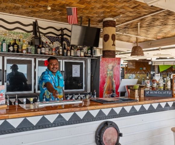 Bartender in blue floral shirt and red flower serves a tropical drink at a festive beach bar. Bottles line the counter; an American flag is visible above.
