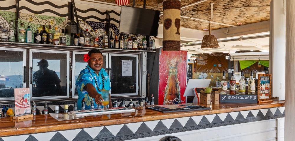 Bartender in blue floral shirt and red flower serves a tropical drink at a festive beach bar. Bottles line the counter; an American flag is visible above.