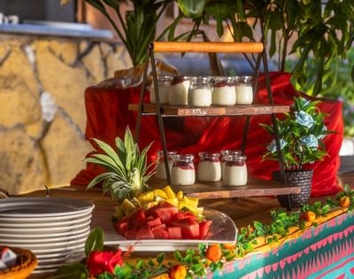 A vibrant buffet table with layered yogurt jars, a plate of fresh fruit slices like watermelon and pineapple, and decorative plants, exuding a festive ambiance.