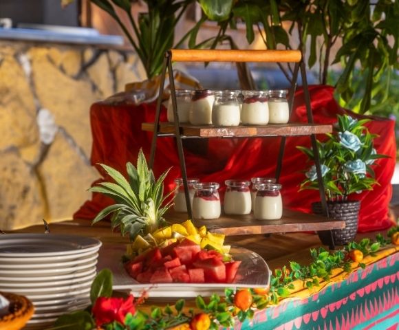 A vibrant buffet table with layered yogurt jars, a plate of fresh fruit slices like watermelon and pineapple, and decorative plants, exuding a festive ambiance.