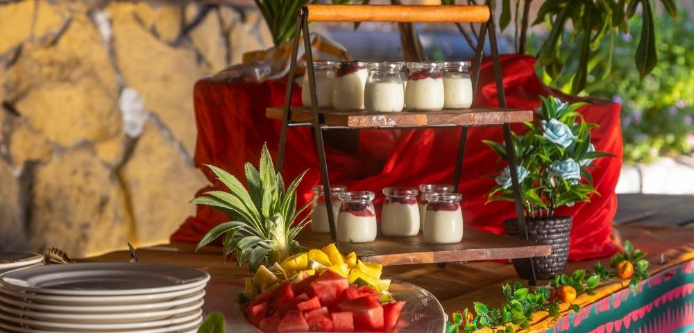A vibrant buffet table with layered yogurt jars, a plate of fresh fruit slices like watermelon and pineapple, and decorative plants, exuding a festive ambiance.