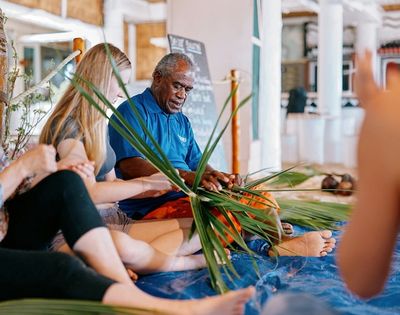A group seated on a blue mat weaves palm leaves, guided by an older man in a blue shirt, indoors. The scene exudes focus and collaboration.