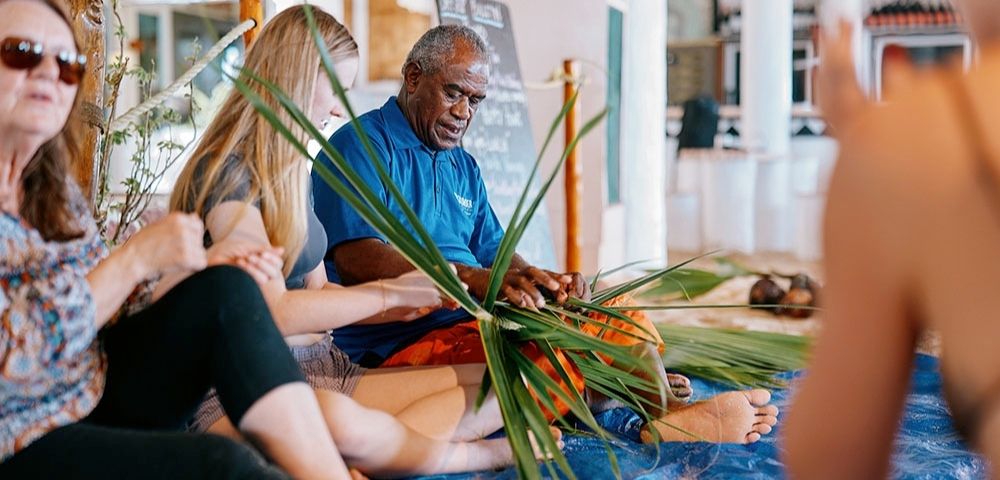 A group seated on a blue mat weaves palm leaves, guided by an older man in a blue shirt, indoors. The scene exudes focus and collaboration.