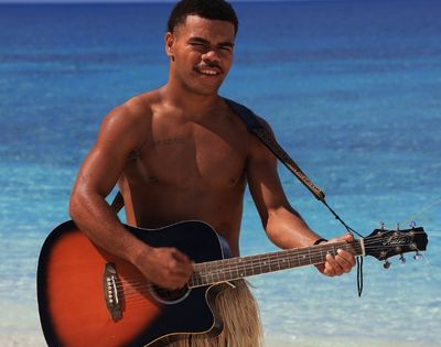 A man plays an acoustic guitar on a tropical beach, wearing a grass skirt. The serene ocean and blue sky create a relaxed, joyful atmosphere.