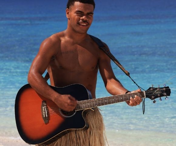 A man plays an acoustic guitar on a tropical beach, wearing a grass skirt. The serene ocean and blue sky create a relaxed, joyful atmosphere.