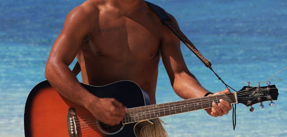 A man plays an acoustic guitar on a tropical beach, wearing a grass skirt. The serene ocean and blue sky create a relaxed, joyful atmosphere.