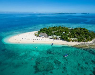 Aerial view of a tropical island surrounded by clear turquoise water, with lush greenery and a sandy beach. Boats are near the shore under a bright blue sky.