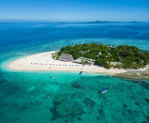 Aerial view of a tropical island surrounded by clear turquoise water, with lush greenery and a sandy beach. Boats are near the shore under a bright blue sky.