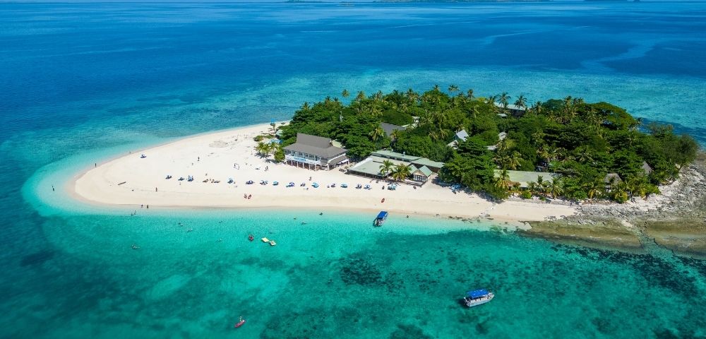 Aerial view of a tropical island surrounded by clear turquoise water, with lush greenery and a sandy beach. Boats are near the shore under a bright blue sky.