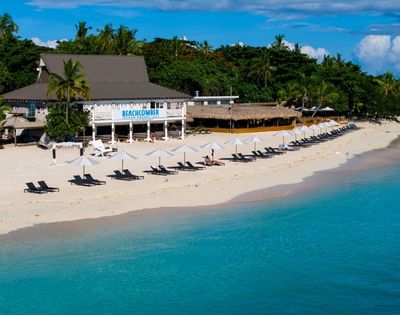 A tropical beach with clear blue waters, lined with white umbrellas and black loungers. A beachfront restaurant is nestled among lush palm trees.