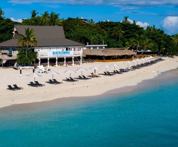 A tropical beach with clear blue waters, lined with white umbrellas and black loungers. A beachfront restaurant is nestled among lush palm trees.