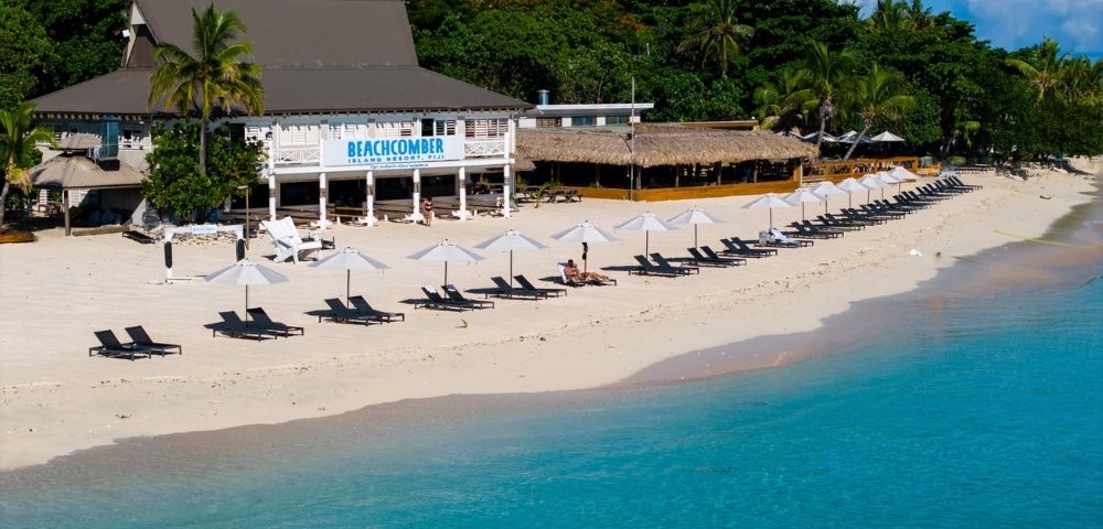 A tropical beach with clear blue waters, lined with white umbrellas and black loungers. A beachfront restaurant is nestled among lush palm trees.