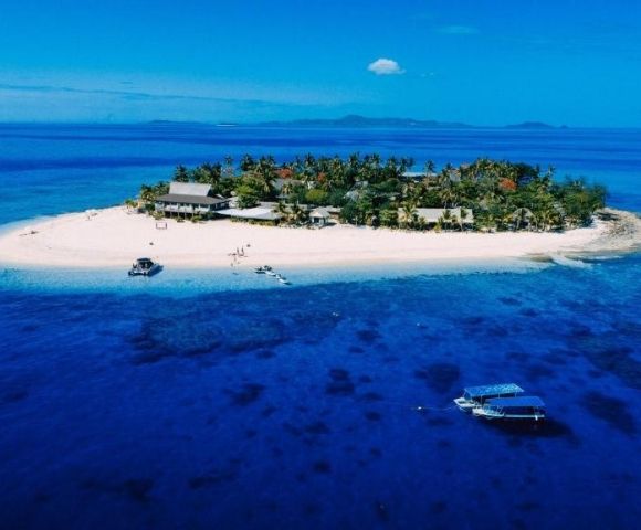 Aerial view of a small tropical island with lush greenery surrounded by clear blue ocean. Boats are anchored nearby, suggesting a tranquil, remote paradise.