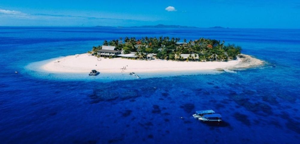 Aerial view of a small tropical island with lush greenery surrounded by clear blue ocean. Boats are anchored nearby, suggesting a tranquil, remote paradise.