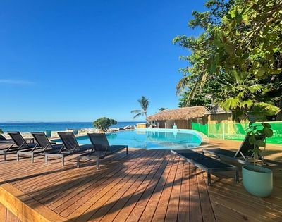 Wooden deck with modern lounge chairs overlooks an infinity pool by the ocean. Lush greenery and clear blue sky create a serene, tropical setting.