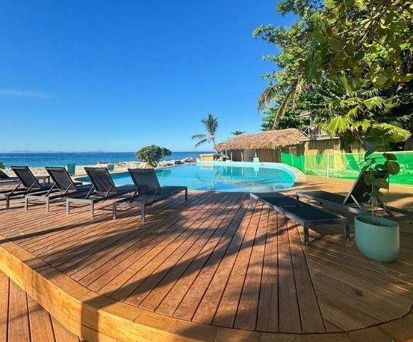 Wooden deck with modern lounge chairs overlooks an infinity pool by the ocean. Lush greenery and clear blue sky create a serene, tropical setting.