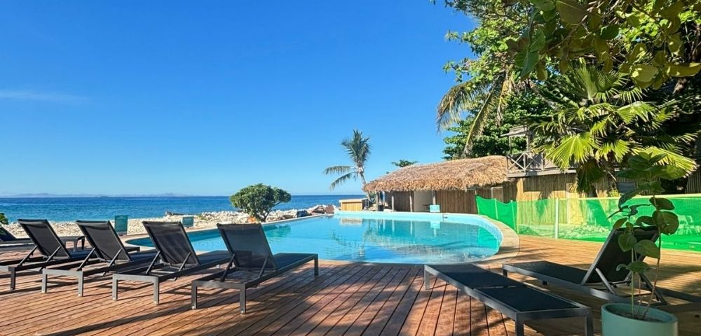 Wooden deck with modern lounge chairs overlooks an infinity pool by the ocean. Lush greenery and clear blue sky create a serene, tropical setting.