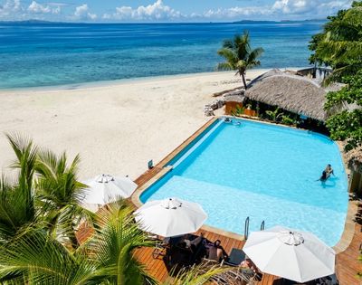 A tranquil beach scene with a clear blue pool beside white sand, palm trees, and turquoise ocean. Sunbathers relax under white umbrellas, conveying serenity.