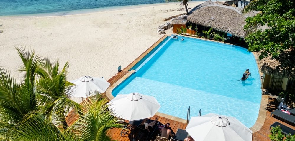 A tranquil beach scene with a clear blue pool beside white sand, palm trees, and turquoise ocean. Sunbathers relax under white umbrellas, conveying serenity.