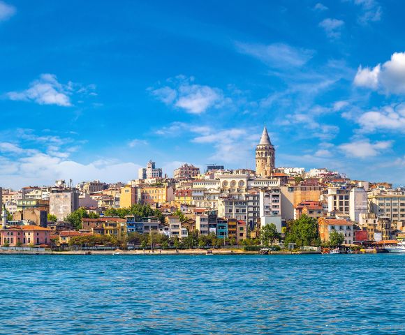 Panoramic view of Istanbul's skyline featuring the Galata Tower amidst colorful buildings under a vibrant blue sky, with the Bosphorus in the foreground.