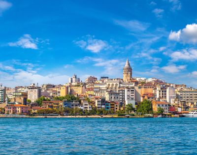 Panoramic view of Istanbul's skyline featuring the Galata Tower amidst colorful buildings under a vibrant blue sky, with the Bosphorus in the foreground.