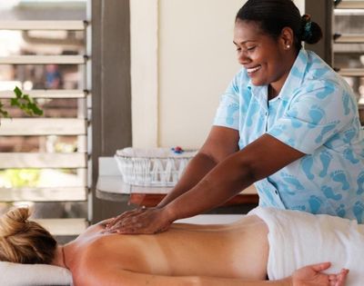 A smiling massage therapist in a blue uniform gives a back massage to a relaxed client in a peaceful, sunlit room, creating a tranquil atmosphere.