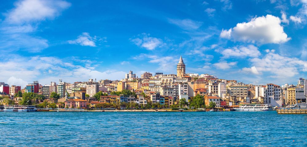 Panoramic view of Istanbul's skyline featuring the Galata Tower amidst colorful buildings under a vibrant blue sky, with the Bosphorus in the foreground.