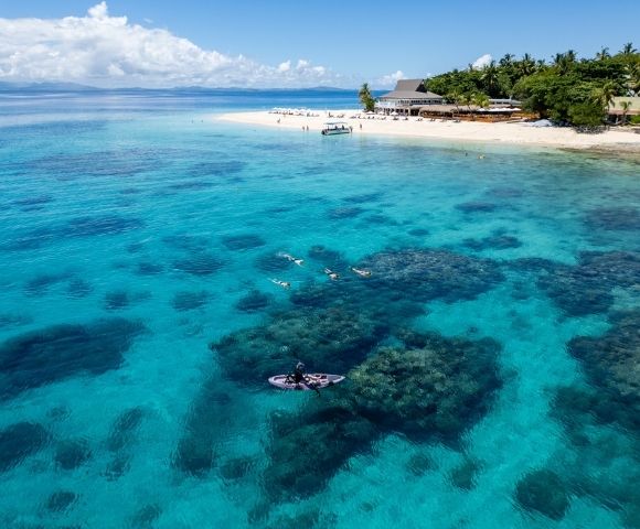 Aerial view of a tropical beach with clear turquoise water and coral reefs. People swim and a boat is docked near a sandy shore lined with lush palm trees.