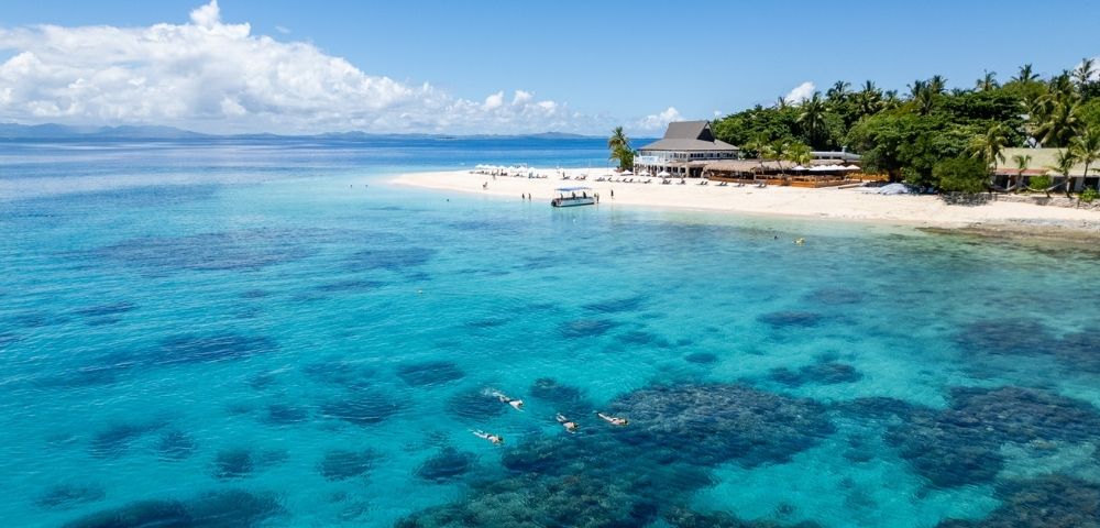 Aerial view of a tropical beach with clear turquoise water and coral reefs. People swim and a boat is docked near a sandy shore lined with lush palm trees.