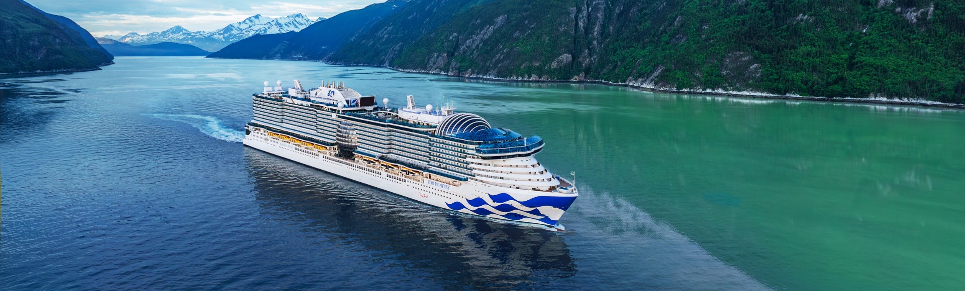 A large cruise ship sails through calm waters surrounded by mountainous landscapes with snow-capped peaks under a partly cloudy sky.
