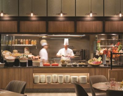 Two chefs wearing white uniforms and tall hats work in a bustling open kitchen. Various ingredients and dishes are artfully displayed, creating a lively, inviting atmosphere.