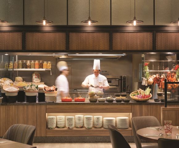 Two chefs wearing white uniforms and tall hats work in a bustling open kitchen. Various ingredients and dishes are artfully displayed, creating a lively, inviting atmosphere.