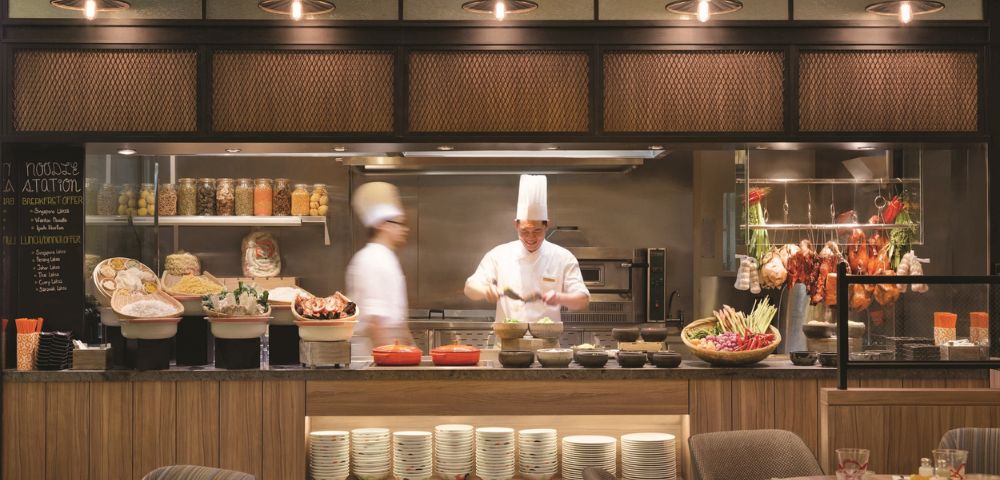 Two chefs wearing white uniforms and tall hats work in a bustling open kitchen. Various ingredients and dishes are artfully displayed, creating a lively, inviting atmosphere.
