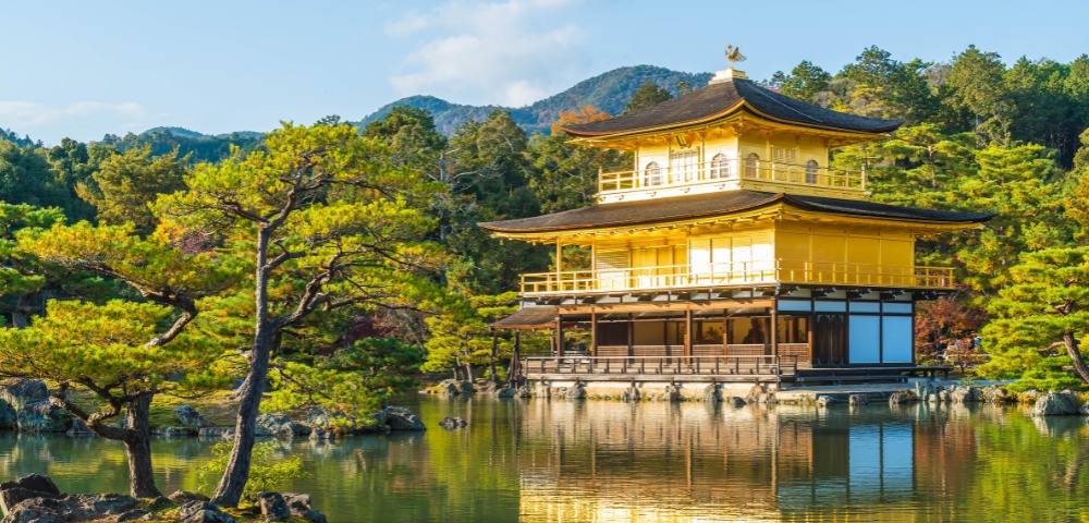Golden pavilion reflecting in a serene pond, surrounded by lush green trees and mountains under a blue sky, creating a tranquil and majestic scene.