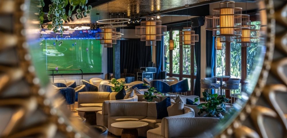 A cozy lounge reflected in a round, ornate mirror, featuring soft beige sofas, hanging pendant lights, and a large TV screen displaying a soccer match.