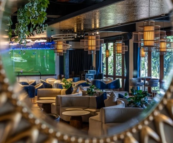 A cozy lounge reflected in a round, ornate mirror, featuring soft beige sofas, hanging pendant lights, and a large TV screen displaying a soccer match.