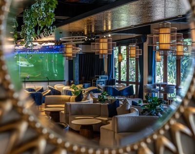 A cozy lounge reflected in a round, ornate mirror, featuring soft beige sofas, hanging pendant lights, and a large TV screen displaying a soccer match.