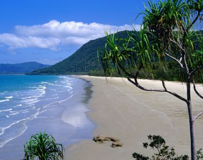 Tropical beach scene with turquoise ocean waves lapping a sandy shore. Green hills and palm trees line the coast under a clear blue sky. Tranquil vibe.